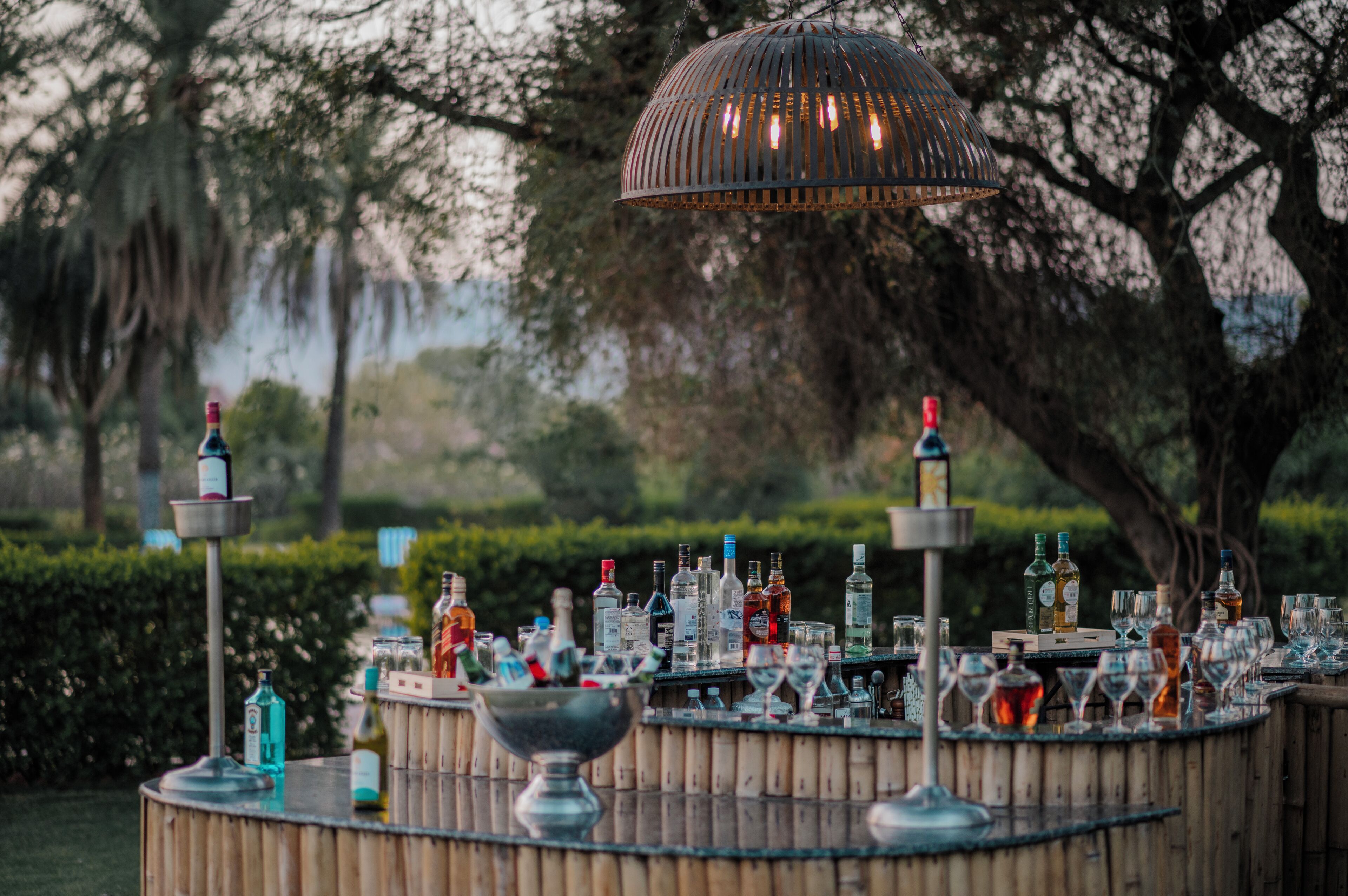 A bar area with a circular wooden table surrounded by bottles and glasses, a hanging light fixture, and a backdrop of trees and greenery.