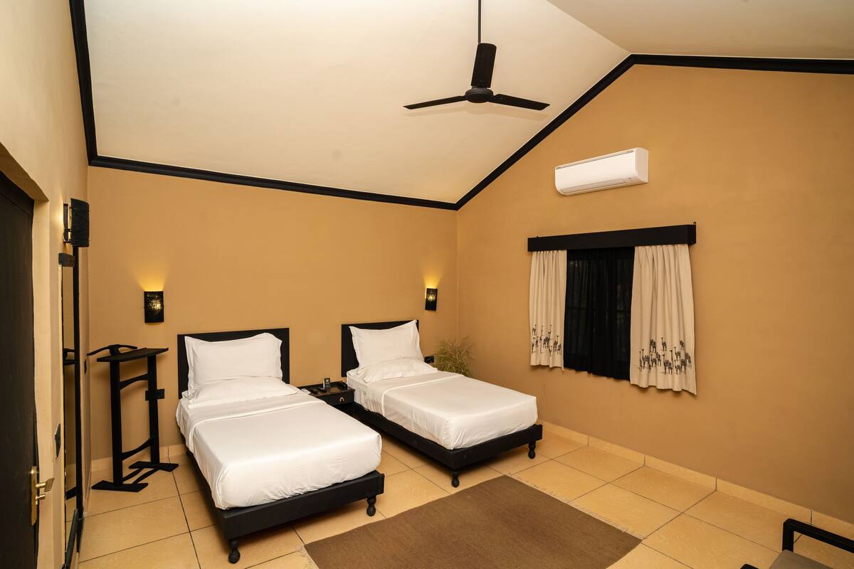 A hotel room with two beds, a ceiling fan, an air conditioning unit, a nightstand, a bedside table, a window with curtains, and a door.