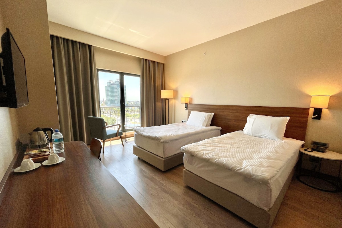 A hotel room with two beds, a desk with a chair, a small table, and a view of the city through large windows.