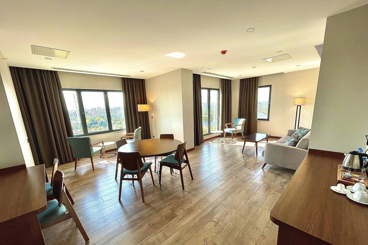 A spacious hotel room with a large dining table, multiple chairs, a sofa, and a small side table with a kettle and cups.