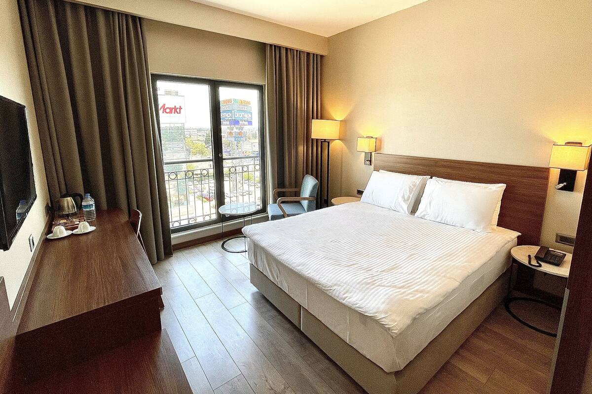 A hotel room with a large bed, a desk with a TV, a chair, a balcony with curtains, and two bedside lamps.