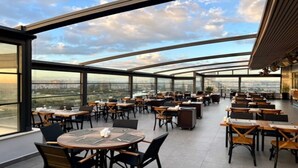 A spacious indoor dining area with tables and chairs, large windows offering a city view, and a curved ceiling structure. - Turkmen Riverside Hotel (Adana)