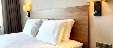 Una habitación de hotel con cama, ropa de cama blanca, lámpara de noche, teléfono y una lámpara de pared.