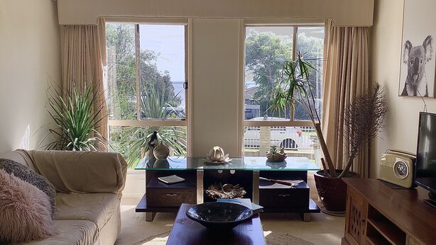 A living room with a sofa, a coffee table, a TV, and a large window with a view of trees and a building. - Maggie’s Nest Come & Rest (Killarney Vale)