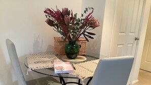 A dining table with a glass top, surrounded by two grey chairs. A woven placemat and a green vase with flowers are on the table. - Maggie’s Nest Come & Rest (Killarney Vale)