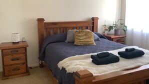 A neatly made bed with a blue blanket, two pillows, and a white throw blanket. There are two bedside tables with drawers and a small vase on one. Three framed artworks are hung on the wall above the bed. - Maggie’s Nest Come & Rest (Killarney Vale)
