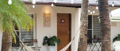 Una hamaca atada entre palmeras, una puerta de madera y una mesa con sillas frente a un edificio.