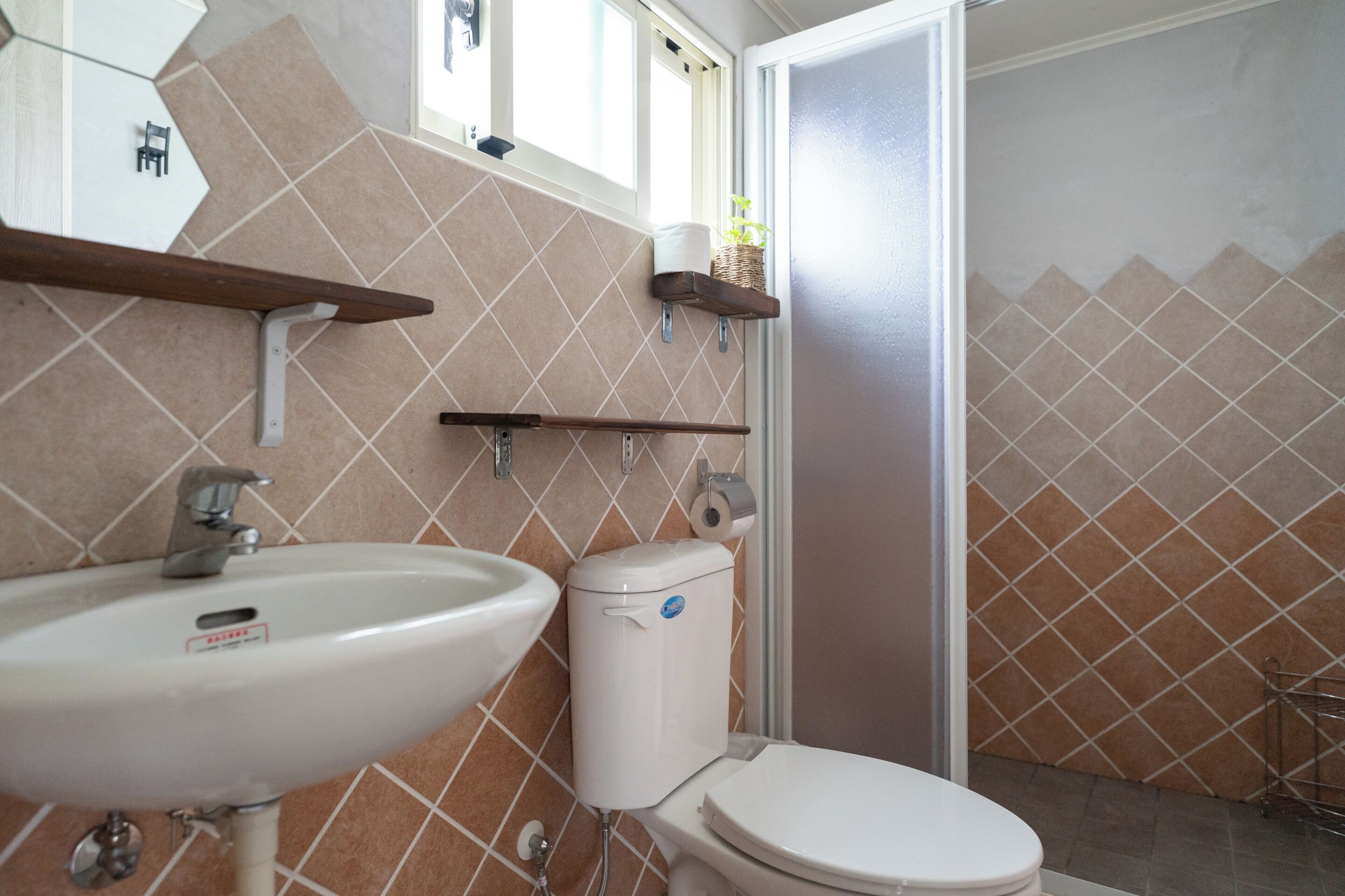 Ein Badezimmer mit einem weißen Waschbecken, einer Toilette und einer Duschkabine. Über Waschbecken und Toilette befinden sich Regale, und oberhalb der Toilette ein Fenster.