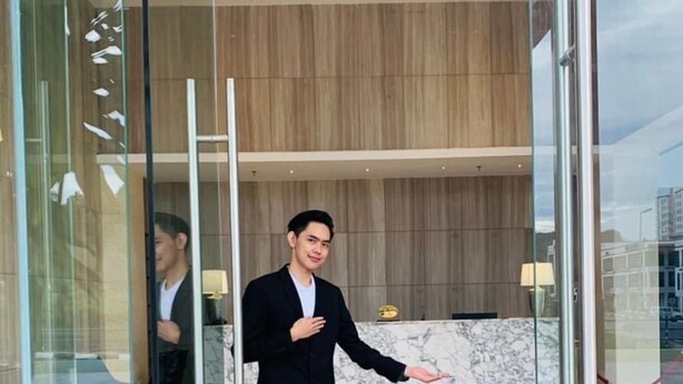 A man in a black suit standing in a lobby with a marble floor, a reception desk, and a wooden wall in the background. - Sentral View Hotel (Bintulu)