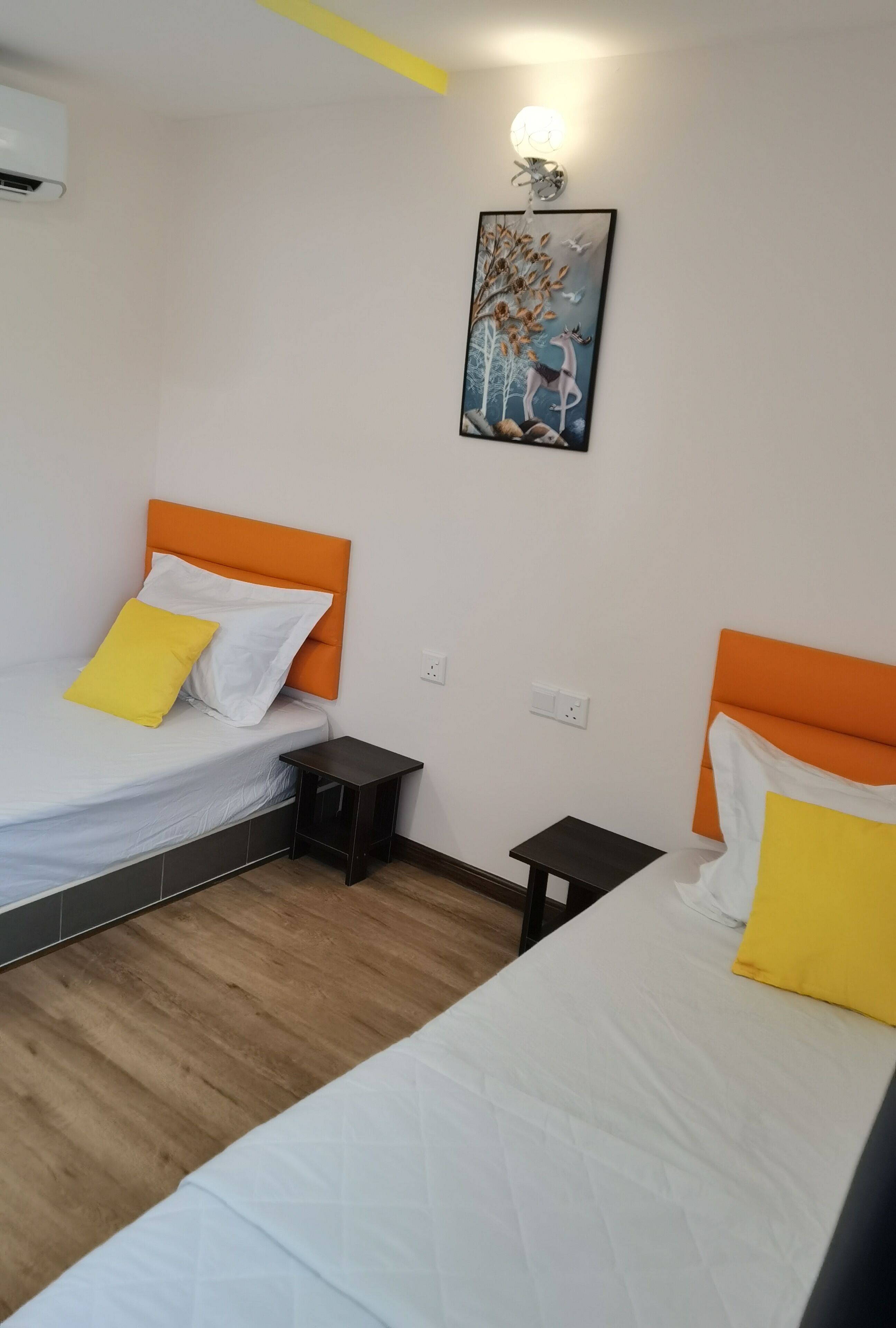 A hotel room with two beds, each with an orange headboard and white bedding. There is a small bedside table on each bed, a wall-mounted air conditioner, and a wall-mounted light fixture.