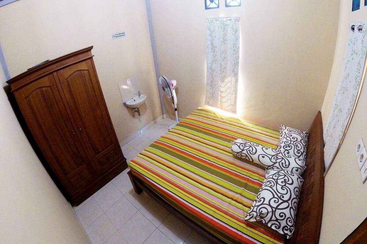 A small bedroom with a bed, a wooden wardrobe, a sink, and a window with curtains.