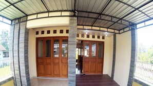 A covered outdoor area with a curved wooden door, a stone pillar, and a tiled floor. - Next Trip Homestay Jogja (Kalasan)