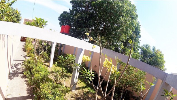 A garden area with various plants and trees, a white fence, and a building in the background. - Next Trip Homestay Jogja (Kalasan)