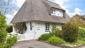A thatched-roof cottage with a white exterior, a small porch, and a garden with various plants and flowers. - Vacation home "Seeräuberhäuschen" (ID 442) (Tating)
