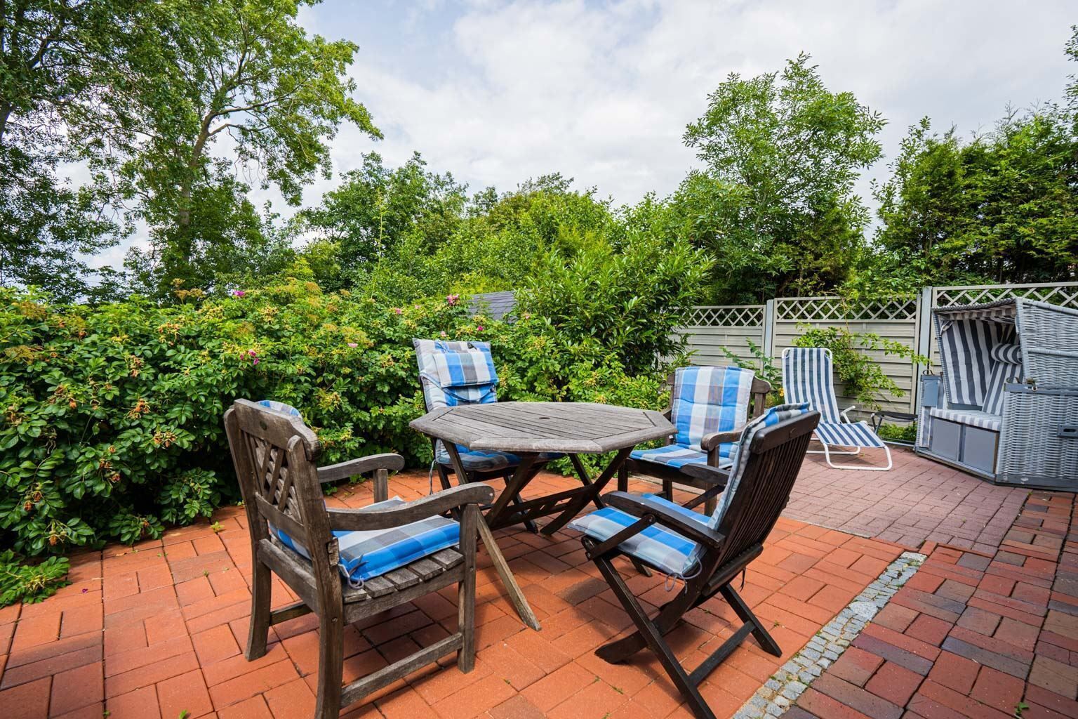 Eine Terrasse im Freien mit einem Holztisch und vier Stühlen, umgeben von üppigem Grün und einem Backsteinboden.