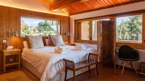 A bedroom with a wooden ceiling, large windows, a bed with white linens, a bedside table, and a chair.