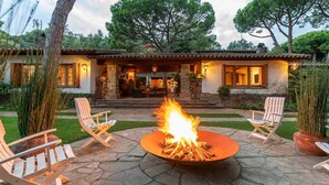 A stone patio with a central fire pit, surrounded by white Adirondack chairs, and a house with a stone facade and warm lighting.