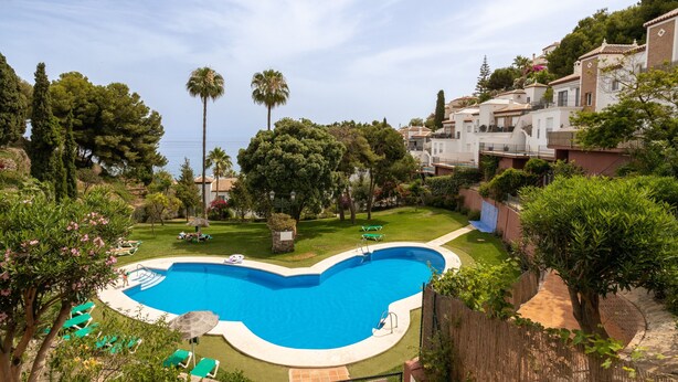 A swimming pool with a curved edge, surrounded by a grassy area with trees and a few lounge chairs. - Apartment 'Casa Sydhavshytten' with Sea View, Wi-Fi and Air Conditioning (Nerja)