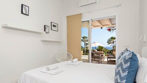 Ett hotellrum med en säng, vita väggar, en vit stol, en balkong med utemöbler och utsikt över havet.