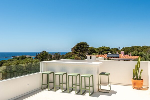 A rooftop terrace with a clear view of the sea, a row of green stools, and a potted cactus.