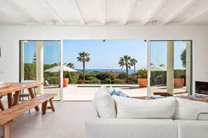 A modern living room with a white sofa, wooden table, and bench, large sliding glass doors leading to a pool and palm trees.