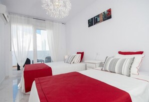 Ein Hotelzimmer mit zwei Betten, einem weißen Nachttisch, einem rot-weißen Kopfkissen, einer weißen Wand und einem Fenster mit durchsichtigen Vorhängen.