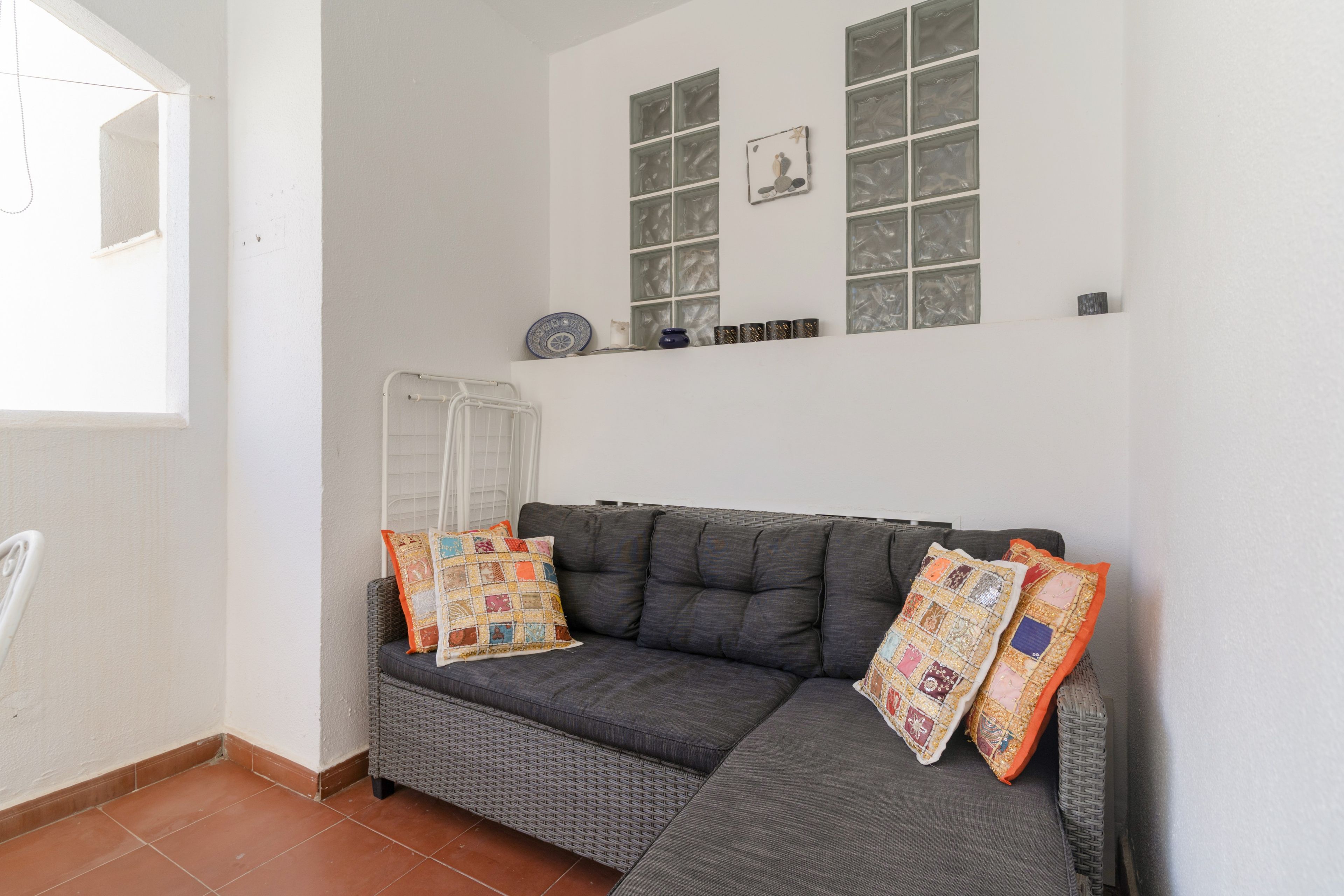 Une pièce avec un canapé gris, deux coussins à motifs, un fauteuil en osier et une fenêtre agrémentée d’objets décoratifs.
