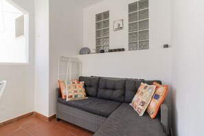 A room with a grey sofa, two patterned cushions, a wicker chair, and a window with decorative items. - Holiday Apartment "Casa Chana" with Terrace, Shared Pool and Jacuzzi (Almunecar)