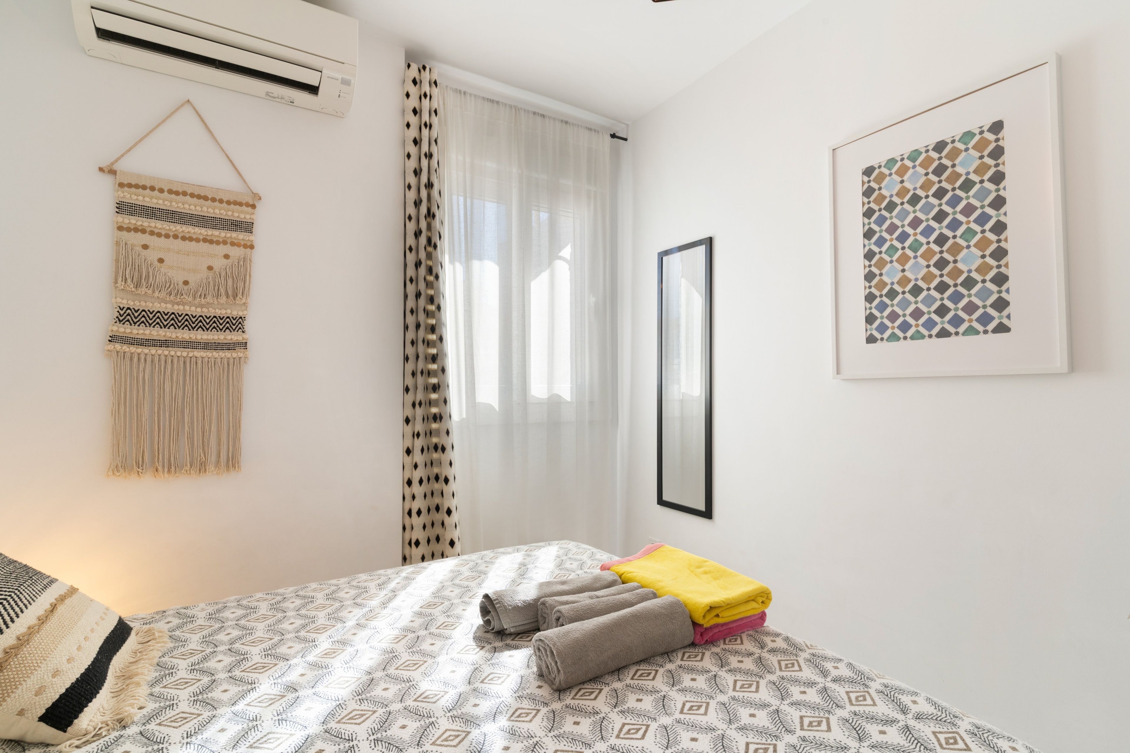 Un dormitorio con cama, aire acondicionado de pared, un tapiz de macramé, un cuadro enmarcado y una ventana con cortinas traslúcidas.