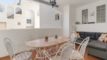 Terraço com mesa redonda e cadeiras brancas, um sofá cinza e uma parede decorativa com objetos emoldurados.