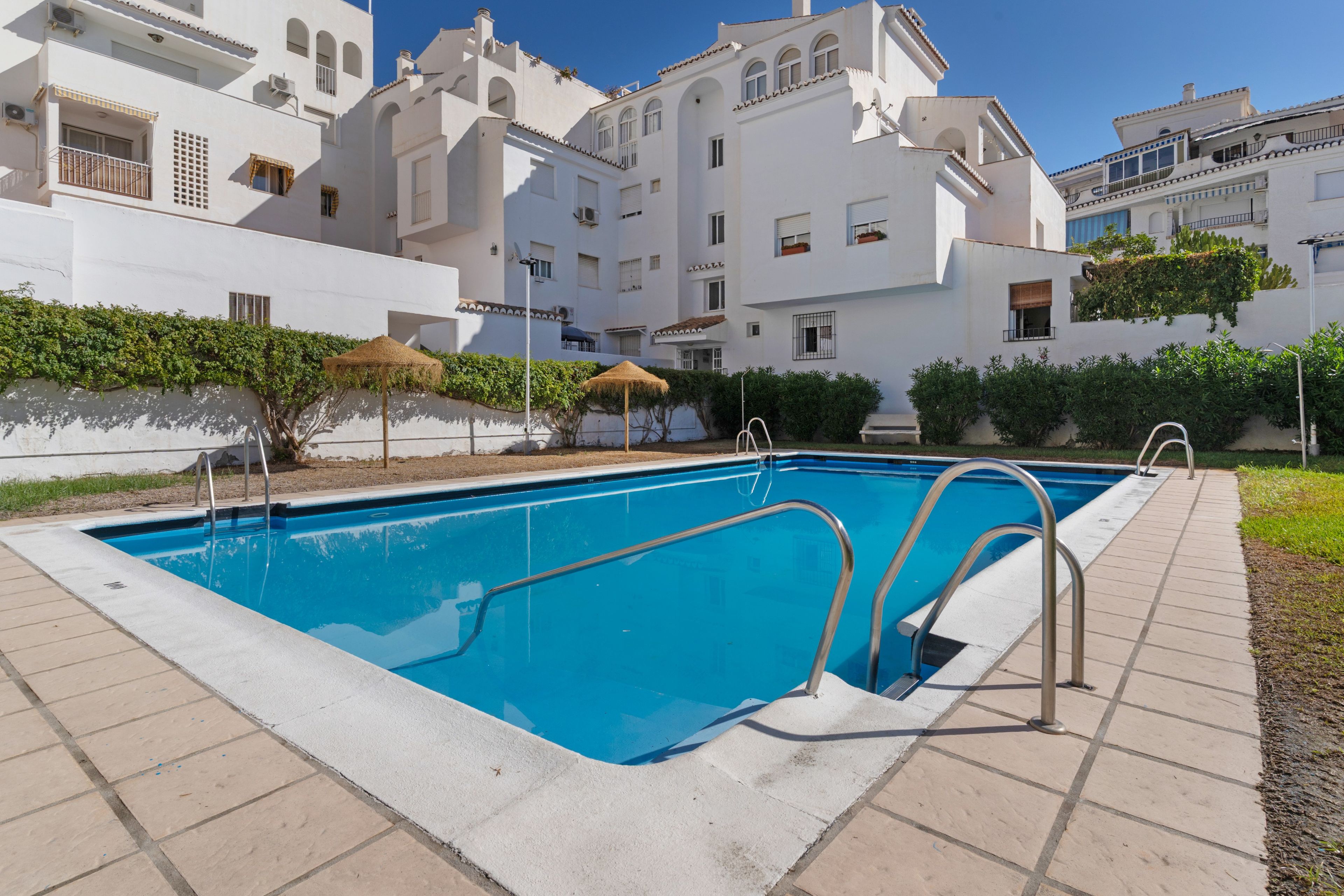 A swimming pool with a tiled deck, surrounded by a white building with balconies and a clear blue sky.