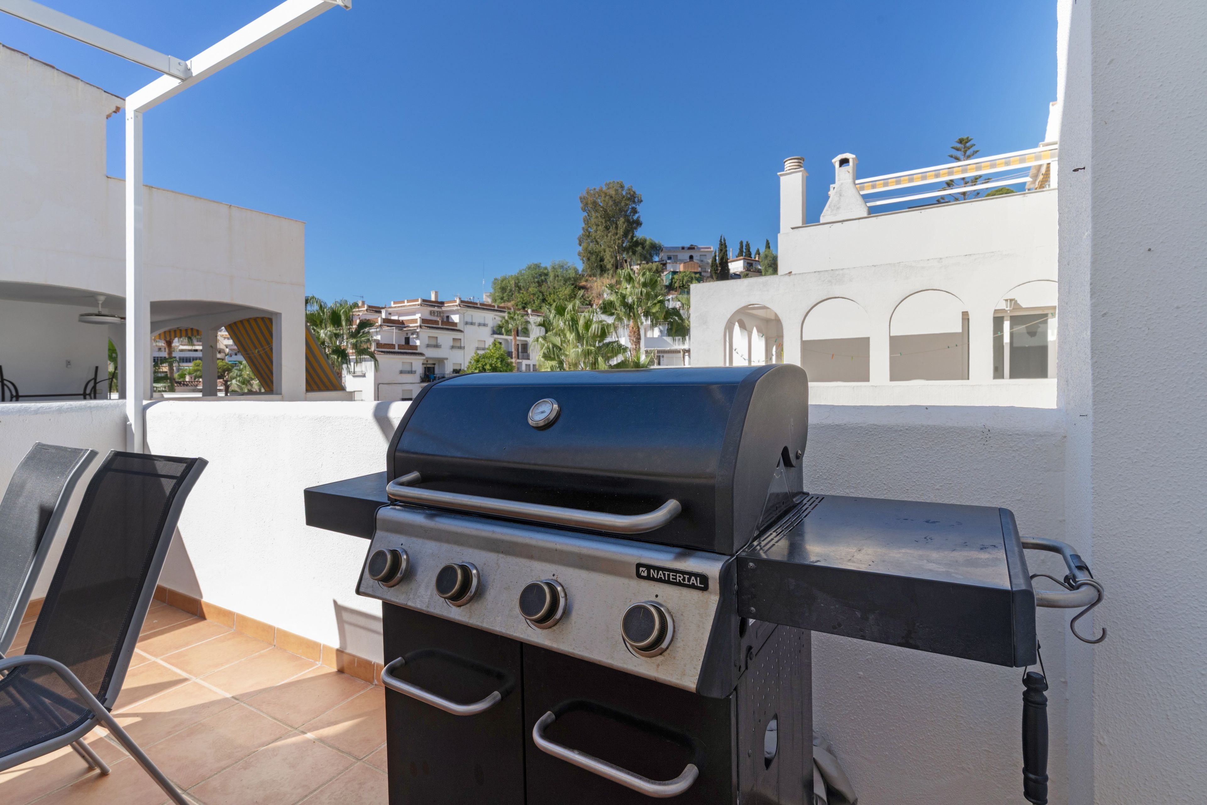 Una parrilla para barbacoa con cuatro quemadores, un quemador lateral y una tapa, ubicada en un patio con una pared blanca y piso de baldosas.