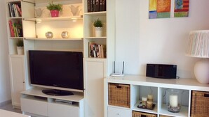 A living room with a flat-screen TV, bookshelves, decorative items, wicker baskets, and a wall-mounted lamp. - Holiday Home 'Vista Marina' with Mountain Views, Garden and Wi-Fi (La Herradura)