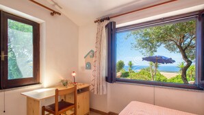 Ein Zimmer mit Bett, Schreibtisch, Stuhl, Fenster mit Blick auf Grünflächen und Strand sowie einer kleinen, hausförmigen Wanddekoration.