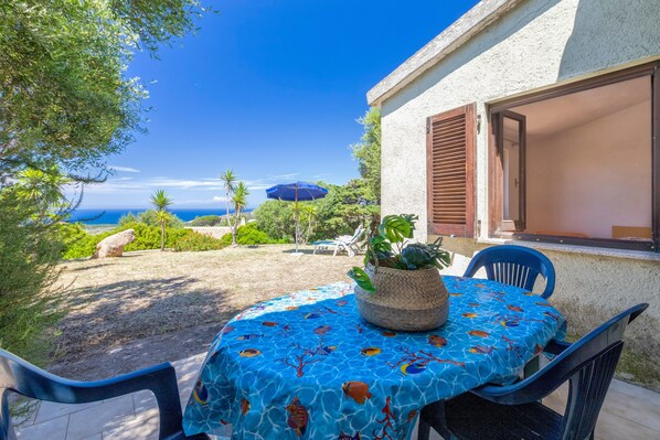 An outdoor patio with a table covered by a blue tablecloth, surrounded by chairs, a potted plant, and a view of the sea.