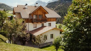 A two-story wooden house with a balcony, surrounded by greenery and mountains.