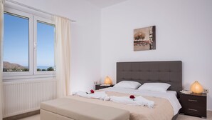 A bedroom with a bed, two bedside tables, a nightstand, a window with curtains, and a view of the sea.