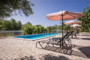 A swimming pool with lounge chairs and umbrellas, surrounded by greenery and a clear sky.