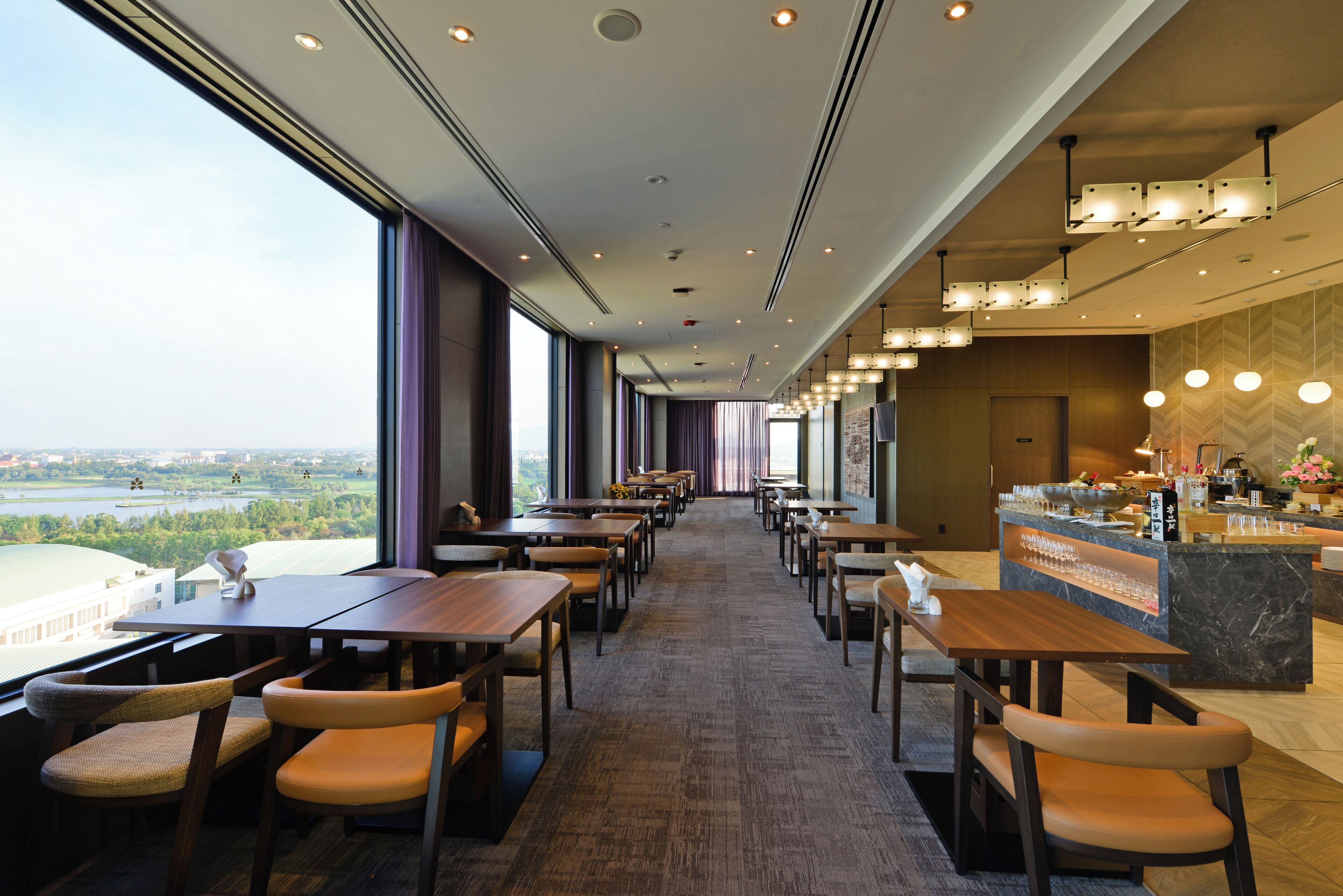 A modern restaurant with a long counter, multiple tables, and chairs. It features large windows with a view of a cityscape and a river.
