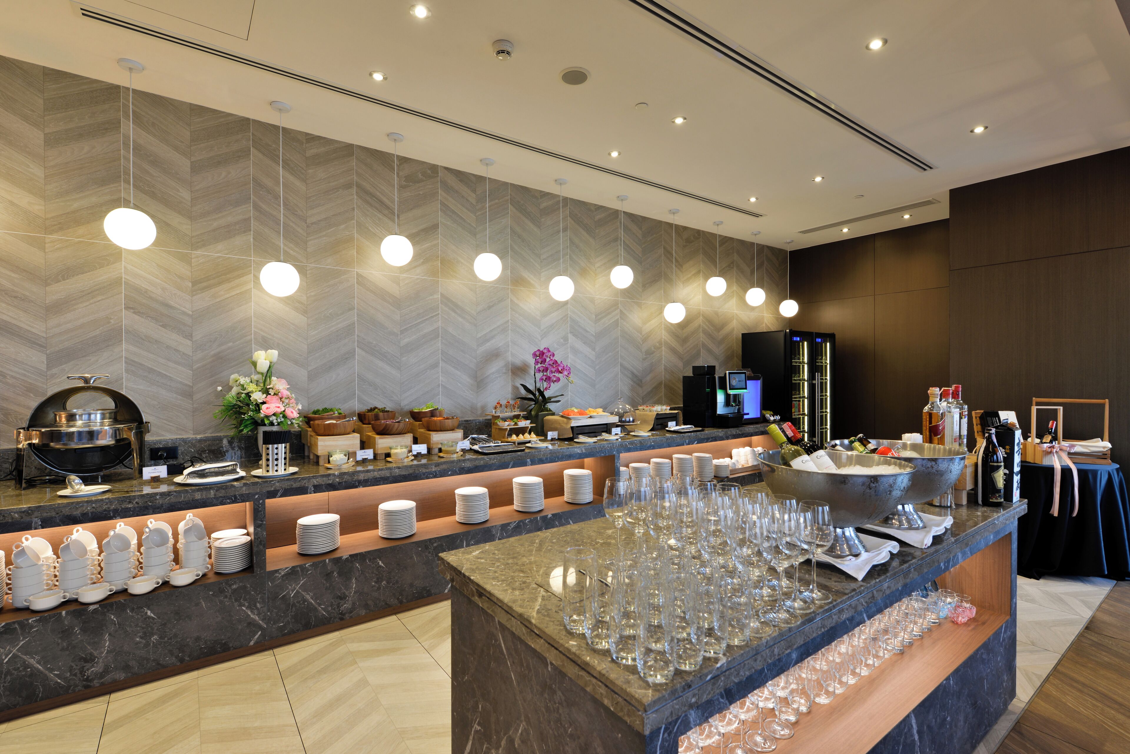 Zona de recepción de un hotel con mostrador de mármol, bufé con diversas opciones de comida y una barra con botellas y vasos.