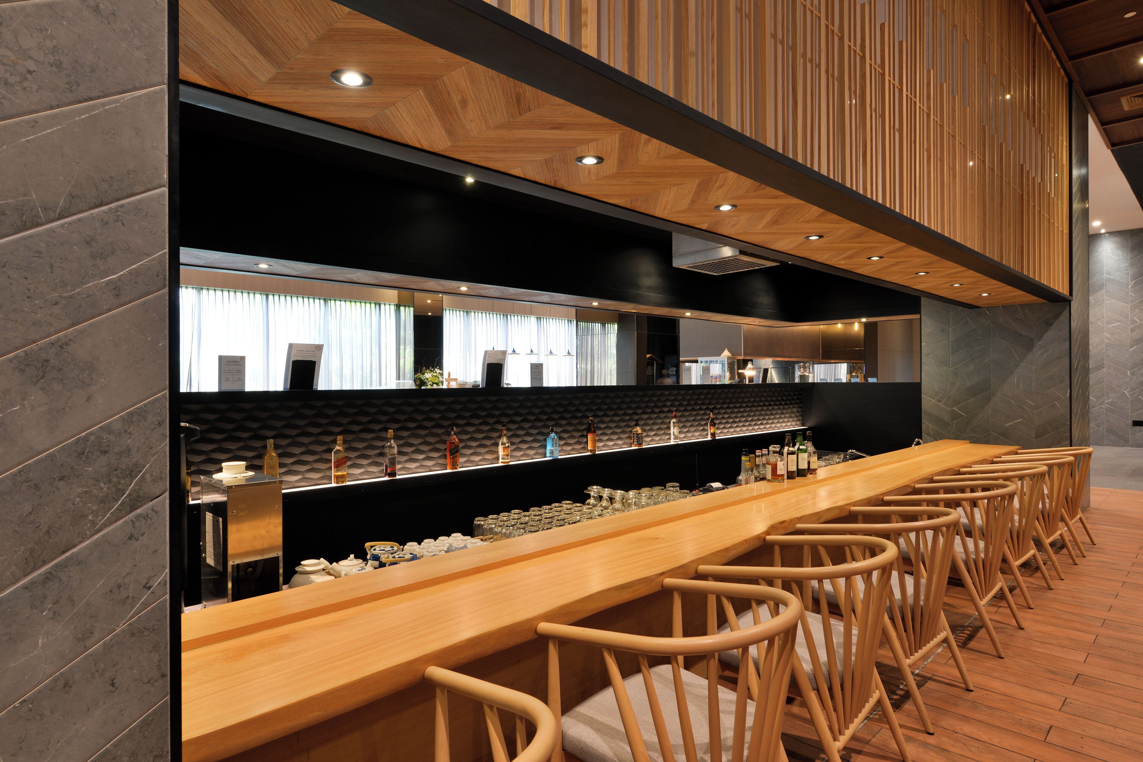 Zona de bar moderna con una larga barra de madera, varios taburetes y un fondo negro con textura.