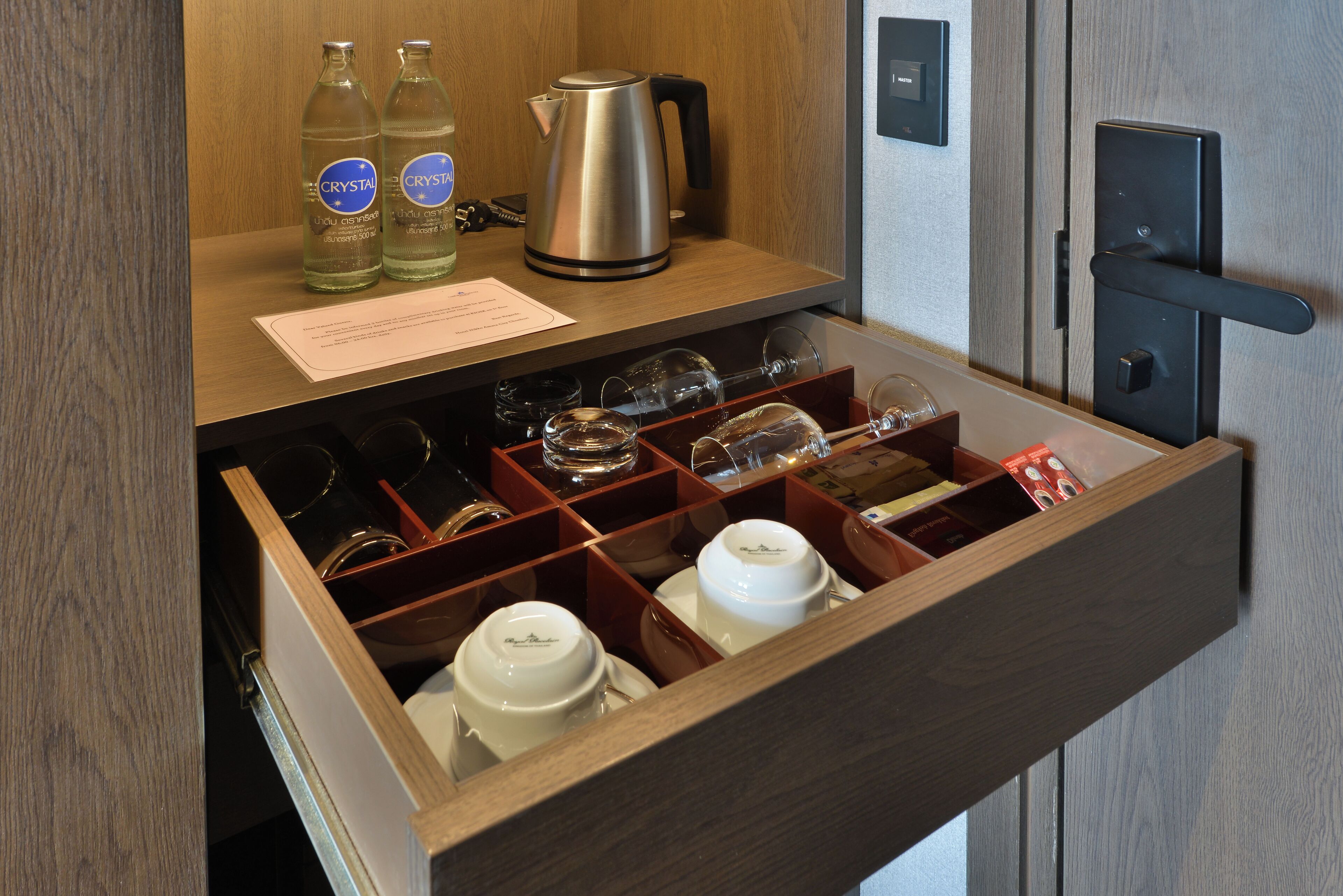 A hotel minibar with neatly organized bottles of water, a kettle, and a variety of glassware and cups.