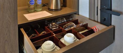 A hotel minibar with neatly organized bottles of water, a kettle, and a variety of glassware and cups.