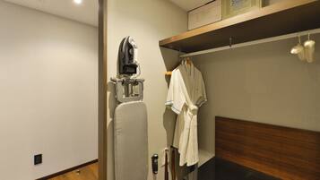 A hotel room with a wooden floor, a bed with a dark headboard, a wall-mounted ironing board, a robe hanging on a rack, and a shelf with a box labeled "GoELECTRON".