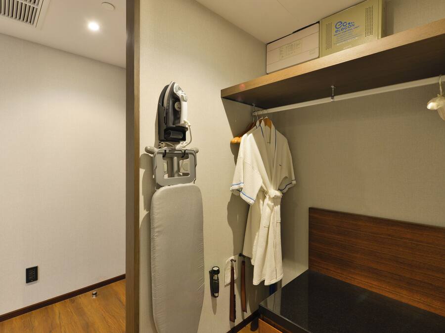 A hotel room with a wooden floor, a bed with a dark headboard, a wall-mounted ironing board, a robe hanging on a rack, and a shelf with a box labeled "GoELECTRON".