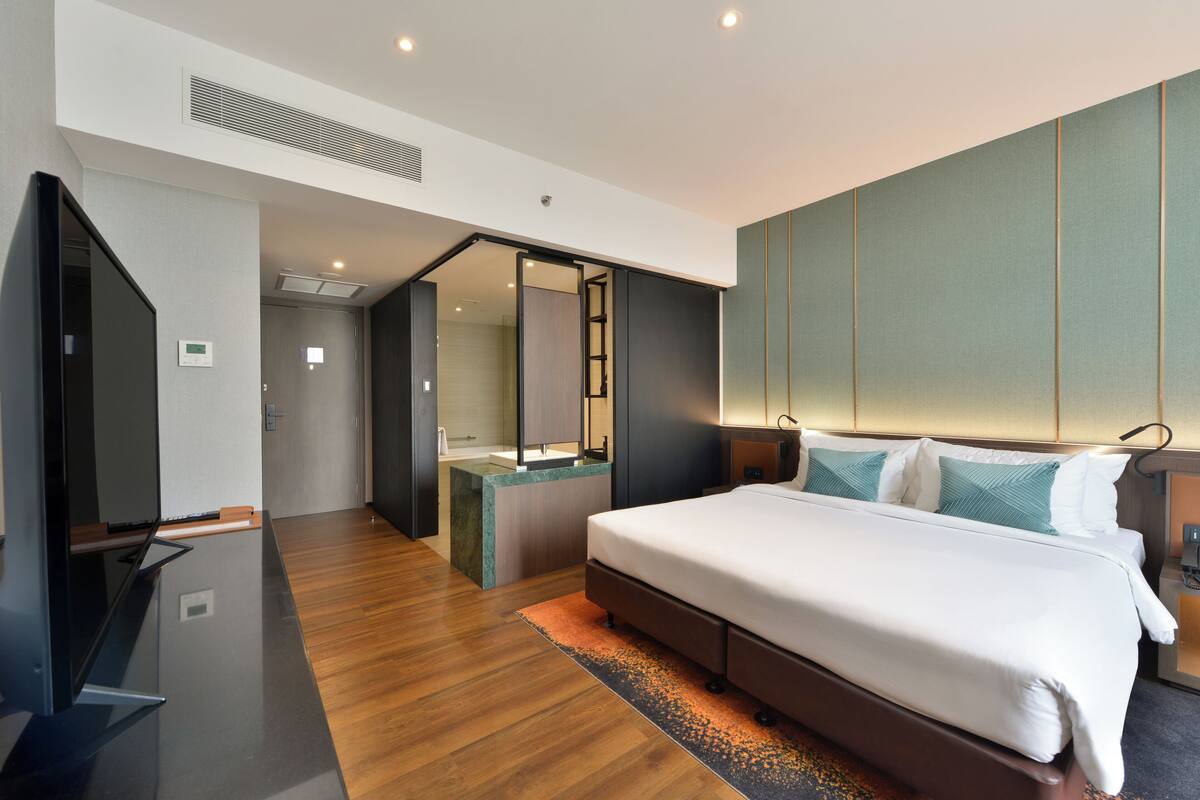 Una habitación de hotel moderna con una cama grande, un televisor de pantalla plana, un baño con lavamanos y espejo, y un vestidor.