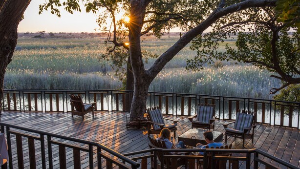 Une terrasse en bois avec des sièges extérieurs et une vue sur un plan d’eau au coucher du soleil.