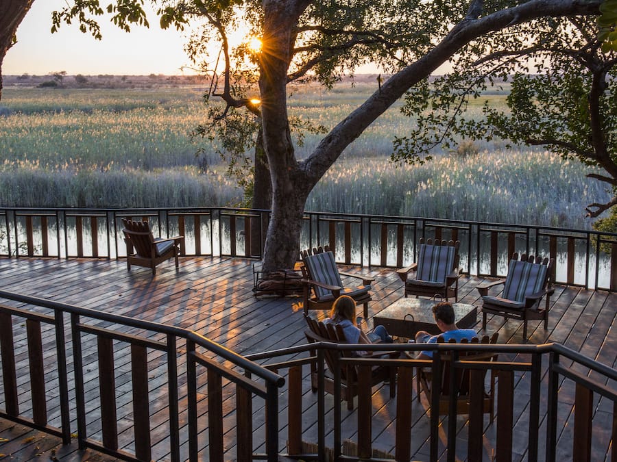 Una terraza de madera con mobiliario exterior y vista a un cuerpo de agua al atardecer.