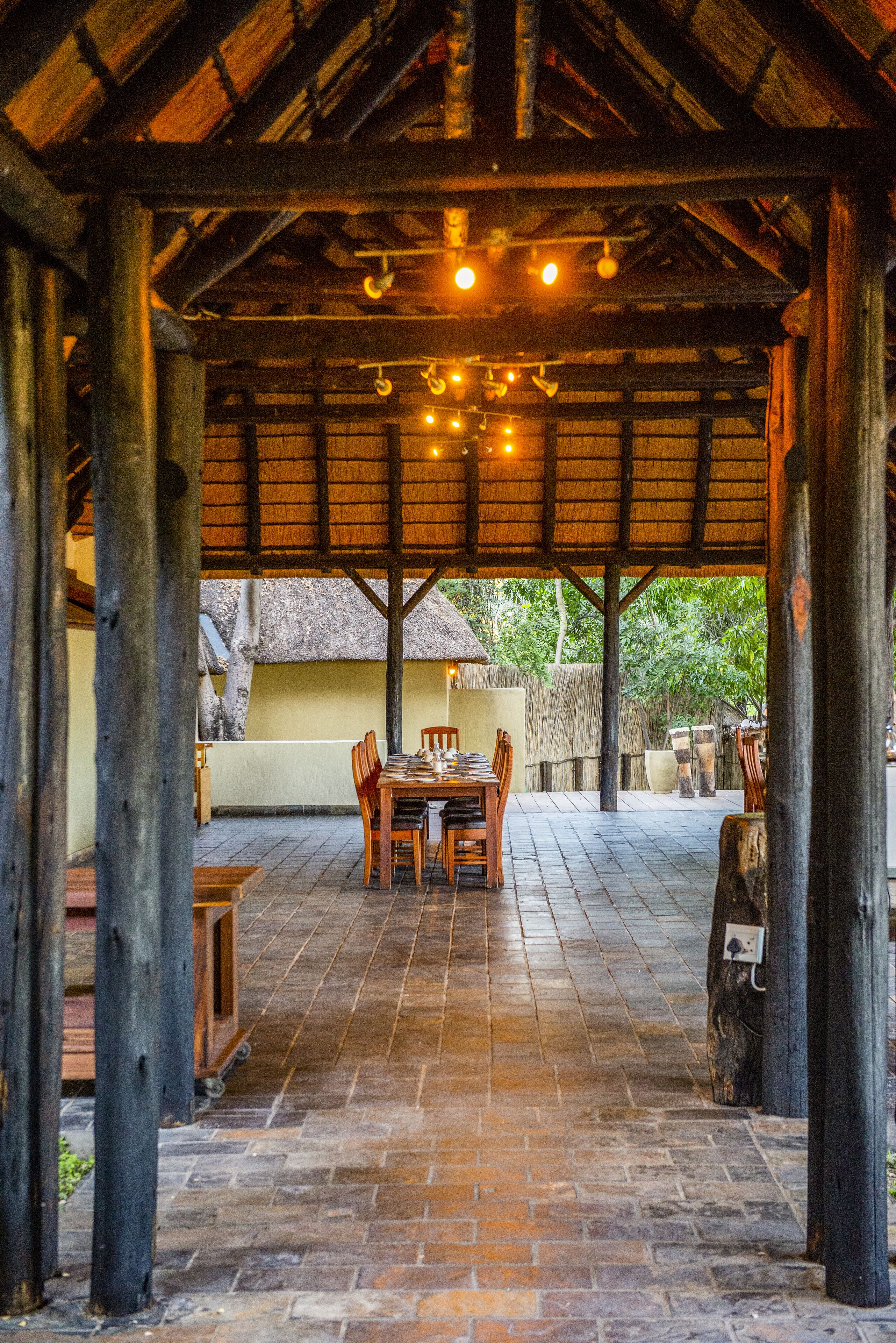 An outdoor covered patio with a wooden table and chairs, surrounded by stone walls and a thatched-roof structure in the background.