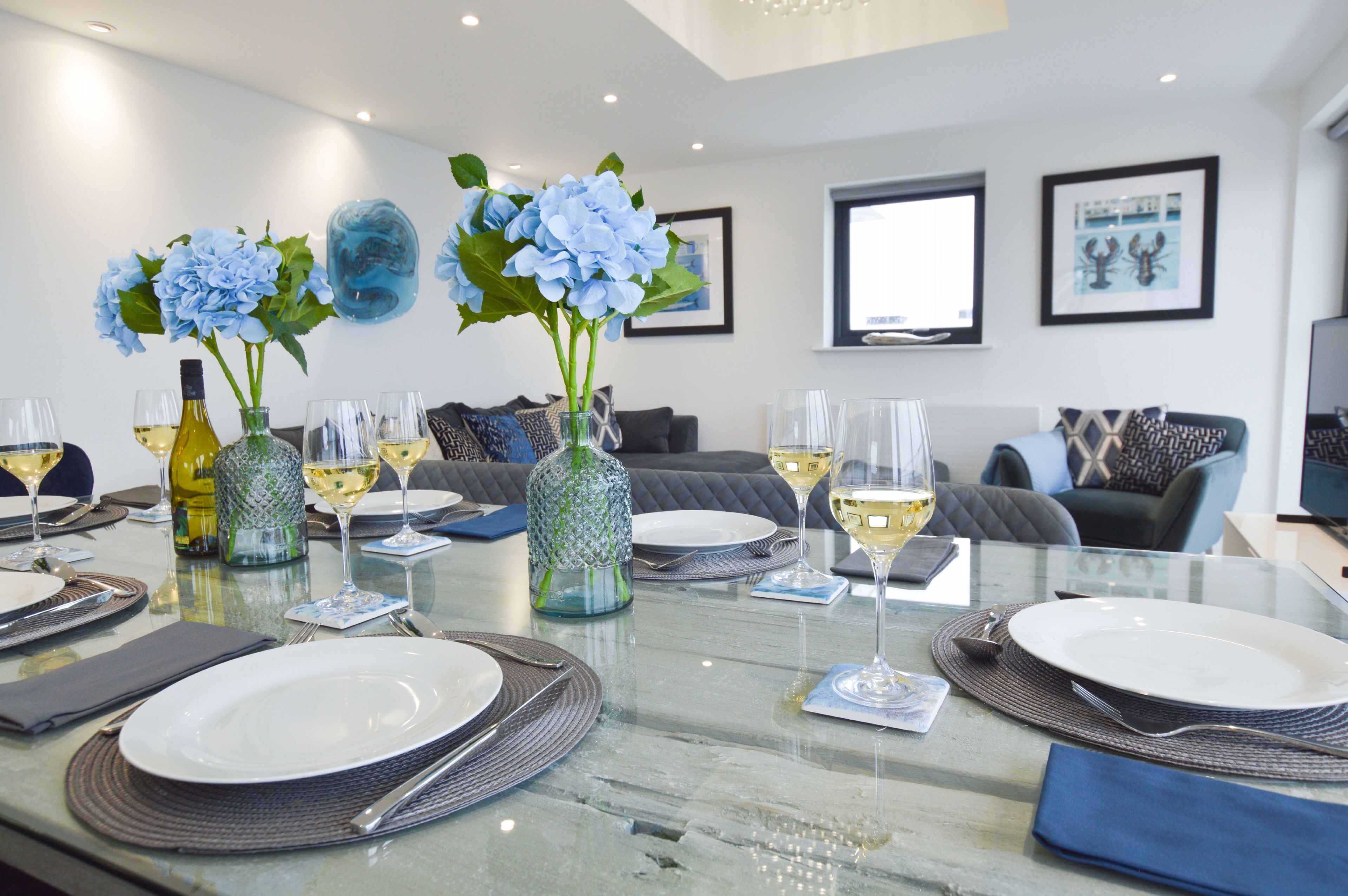 A dining area with a glass table set for a meal, featuring white plates, cutlery, and wine glasses. There are two decorative vases with blue flowers and a bottle of white wine. The room has a modern design with a grey sofa and a framed picture on the wall.